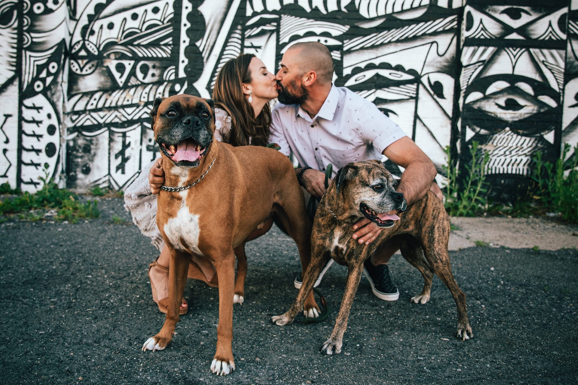 engagement photo of couple kissing with two dogs in front of mural in Eastern Market